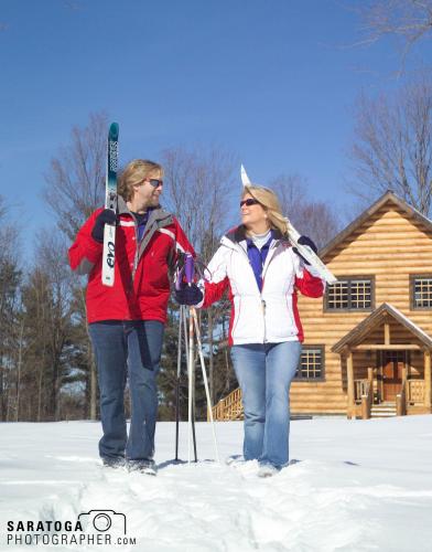 Man and woman in winter attire carrying skis through the snow with log cabin in background