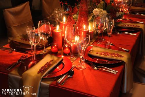 Candlelit view of Christmas holiday table setting with red tablecloth and gold napkins and many crystal glasses